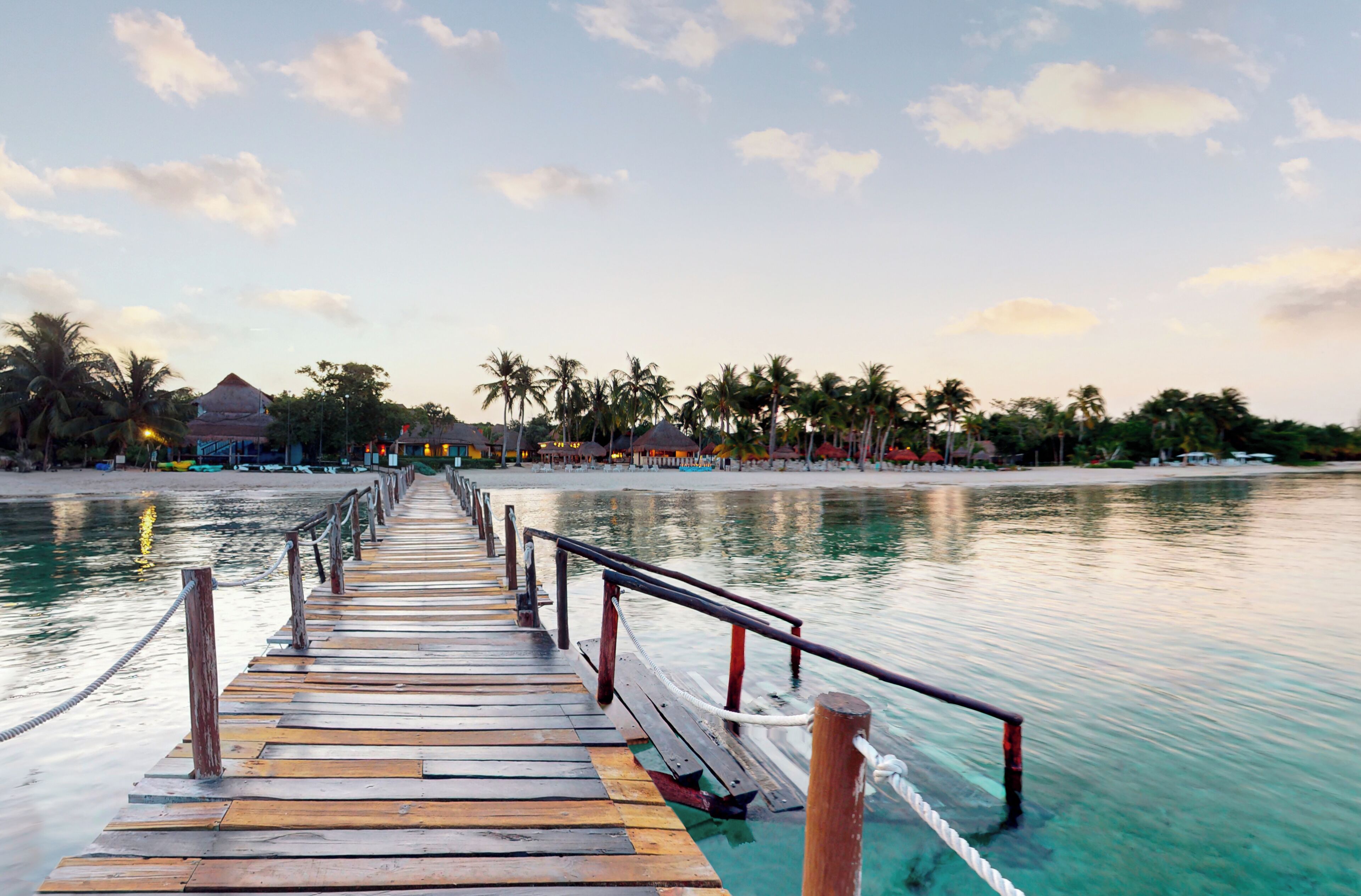 A wooden pier extending into calm waters, leading to a beach with palm trees and beach huts.