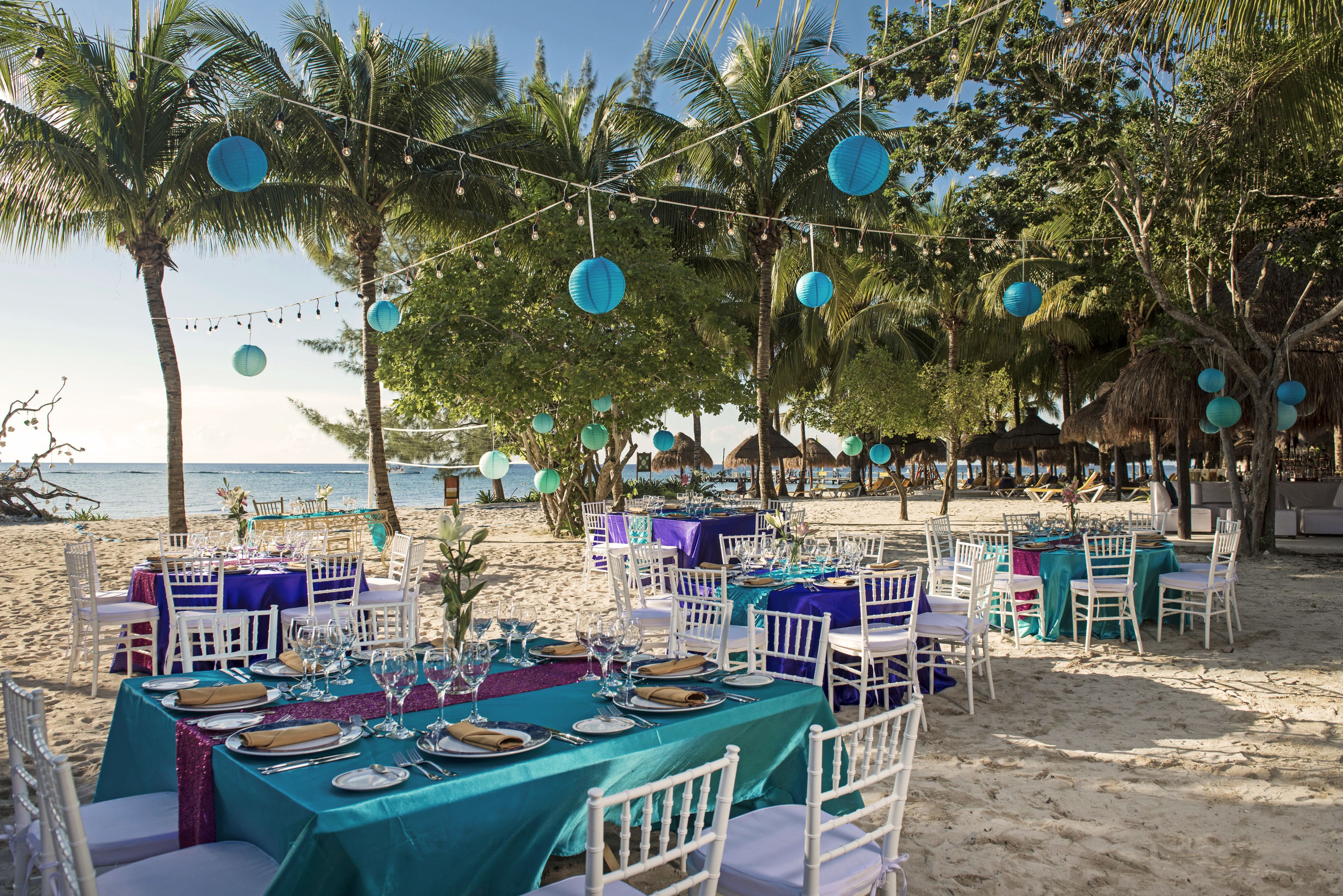 A beachside dining setup with tables and chairs, string lights, and palm trees.