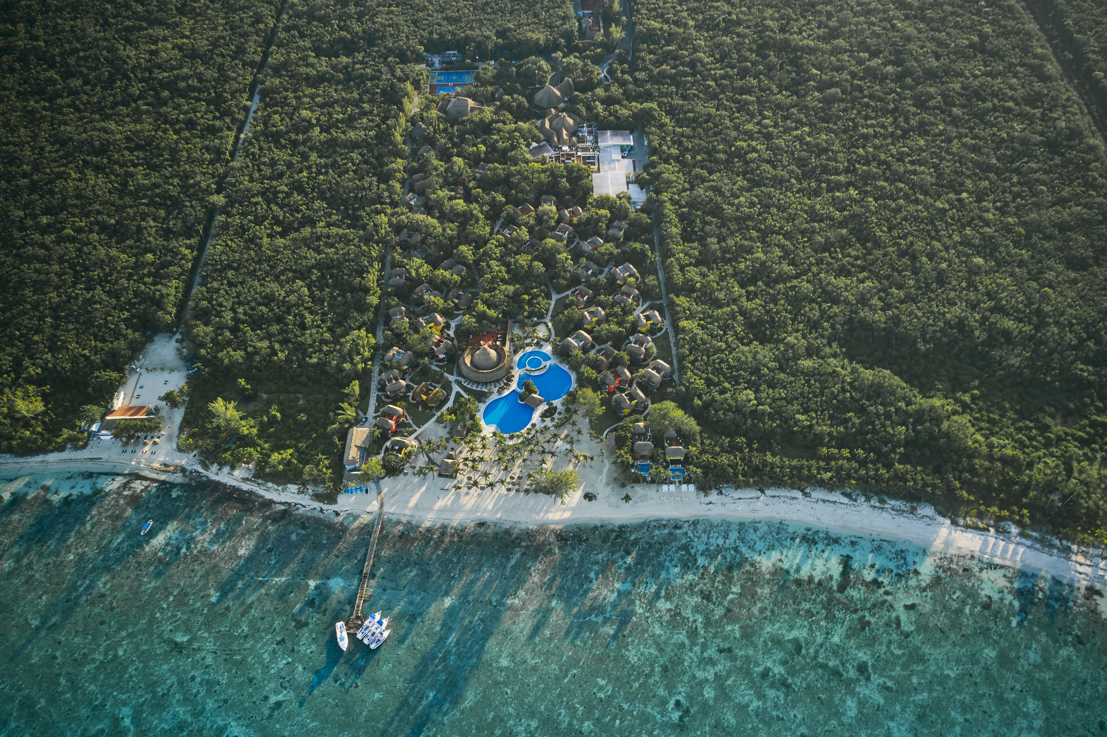 An aerial view of a resort with multiple bungalow-style accommodations, a central swimming pool, and a beach area.