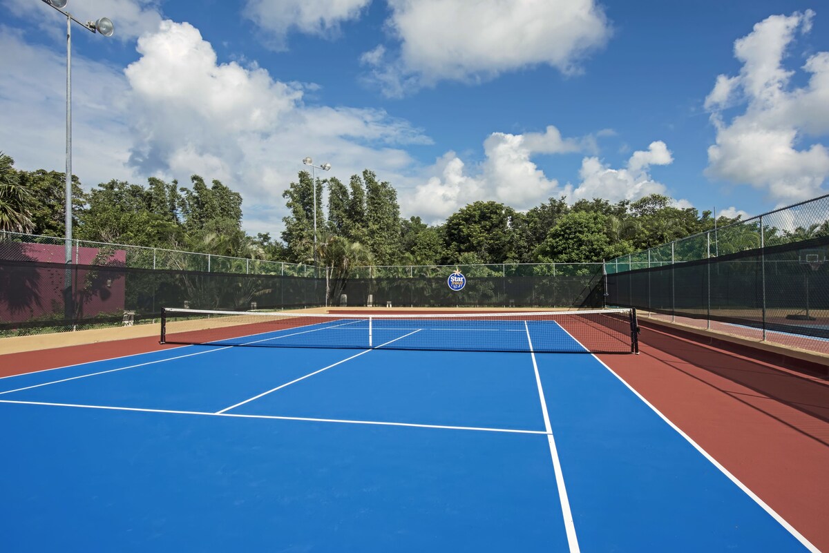 Play a game of tennis on the outdoor court surrounded by lush trees and a nearby building.