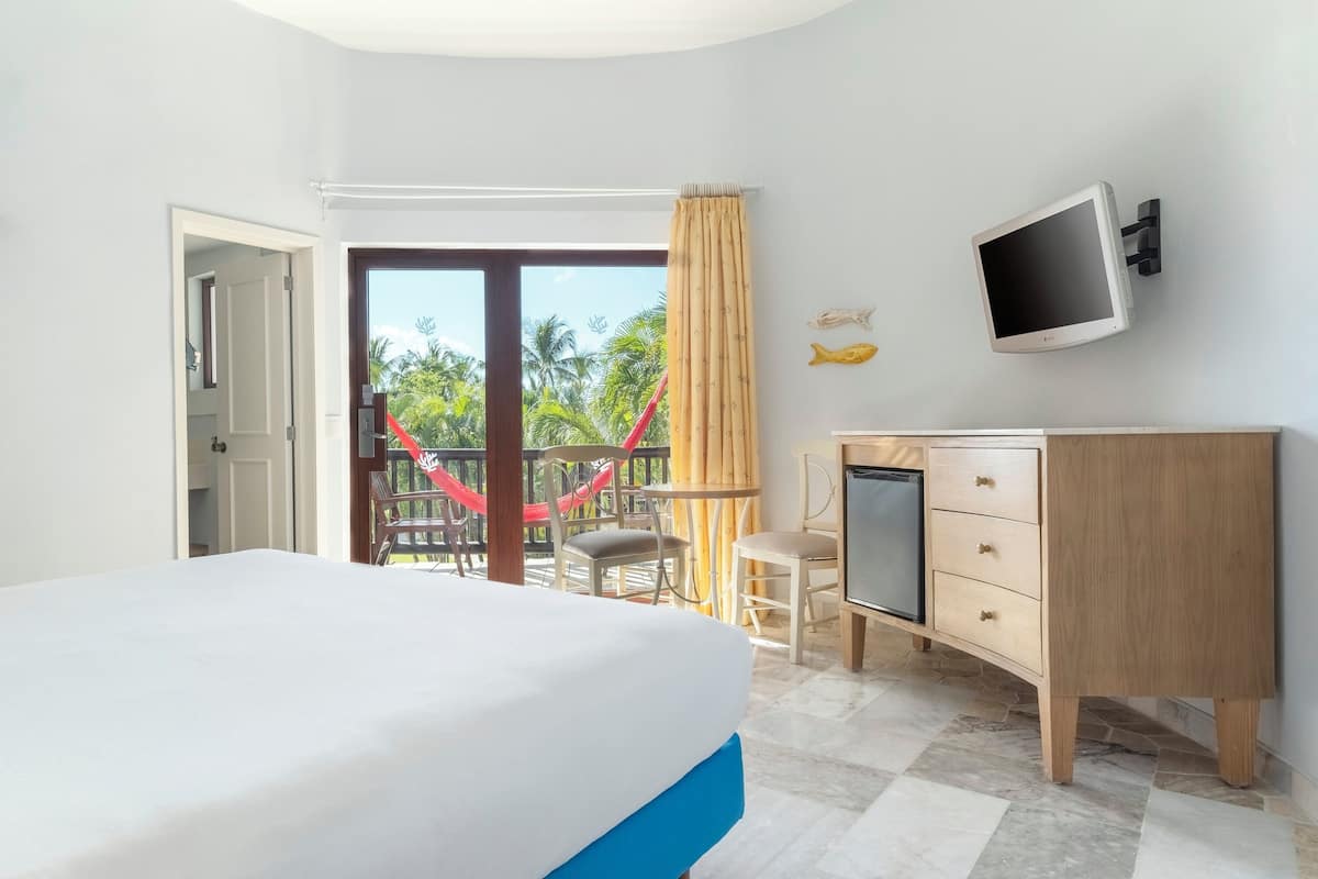 A hotel room with a bed, a desk with a TV, a small fridge, and a view of a balcony with a red swing.