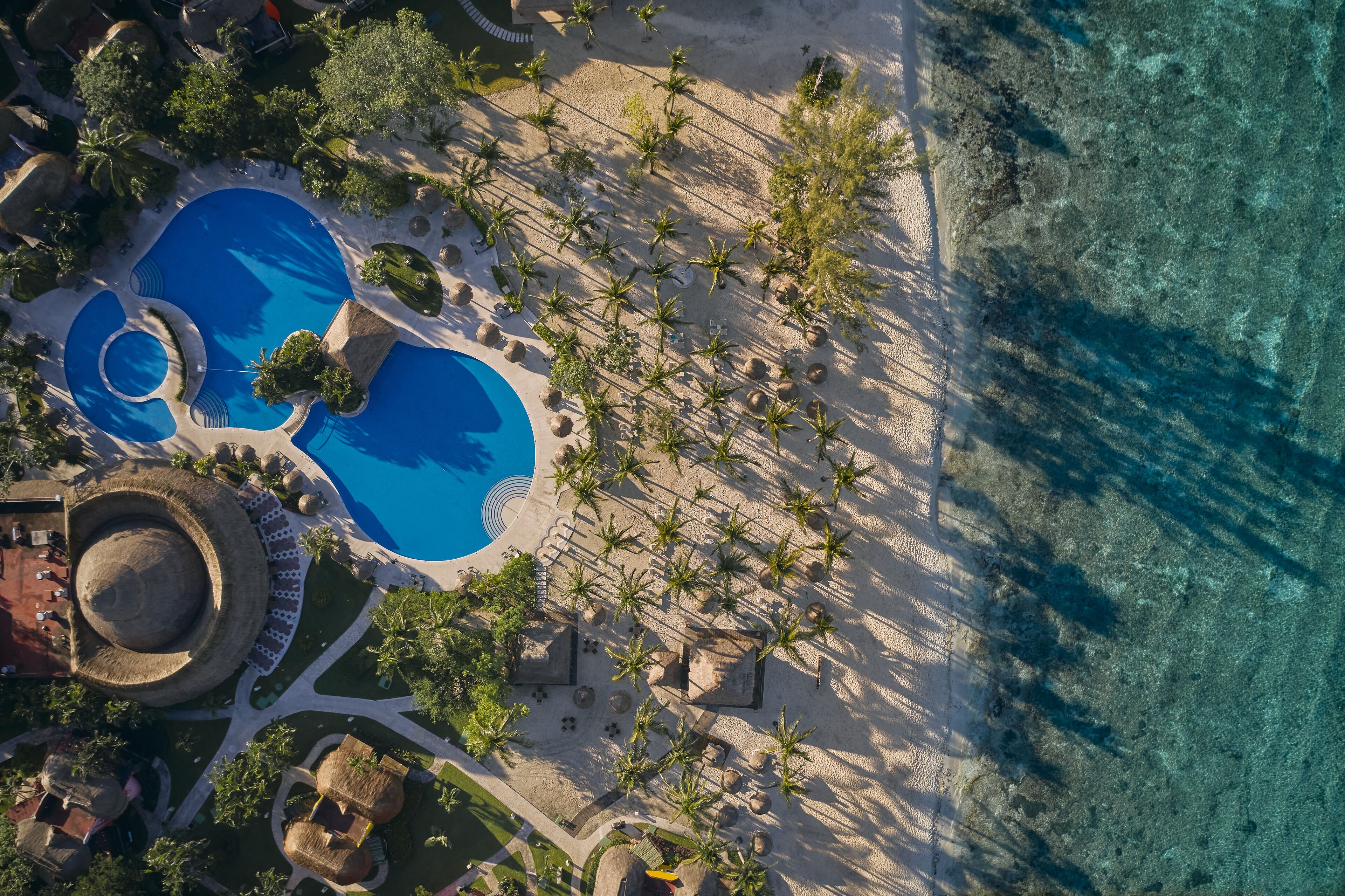 Vista aérea de un complejo turístico con una amplia piscina, palmeras y una zona de playa.