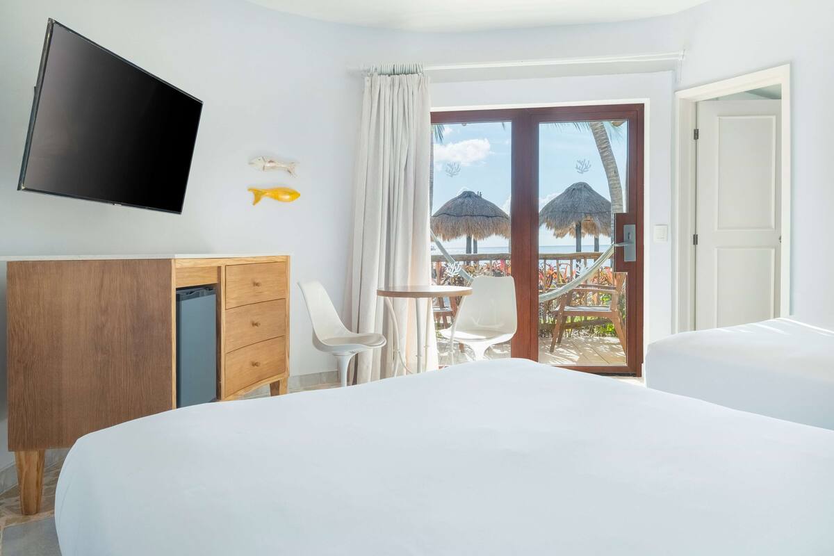 Ein Hotelzimmer mit Bett, Schreibtisch, Stuhl, Fernseher und einem Balkon mit Blick auf Palmen und Strand.