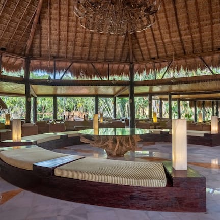 A spacious indoor area with a thatched roof, cushioned seating, and a central wooden table.
