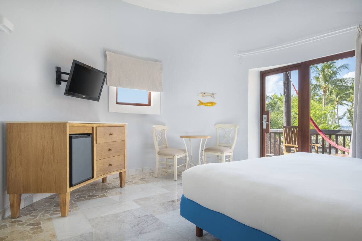 Ein Hotelzimmer mit einem Bett, einem an der Wand befestigten Fernseher, einem Schreibtisch mit zwei Stühlen und Blick auf Palmen durch ein Fenster.