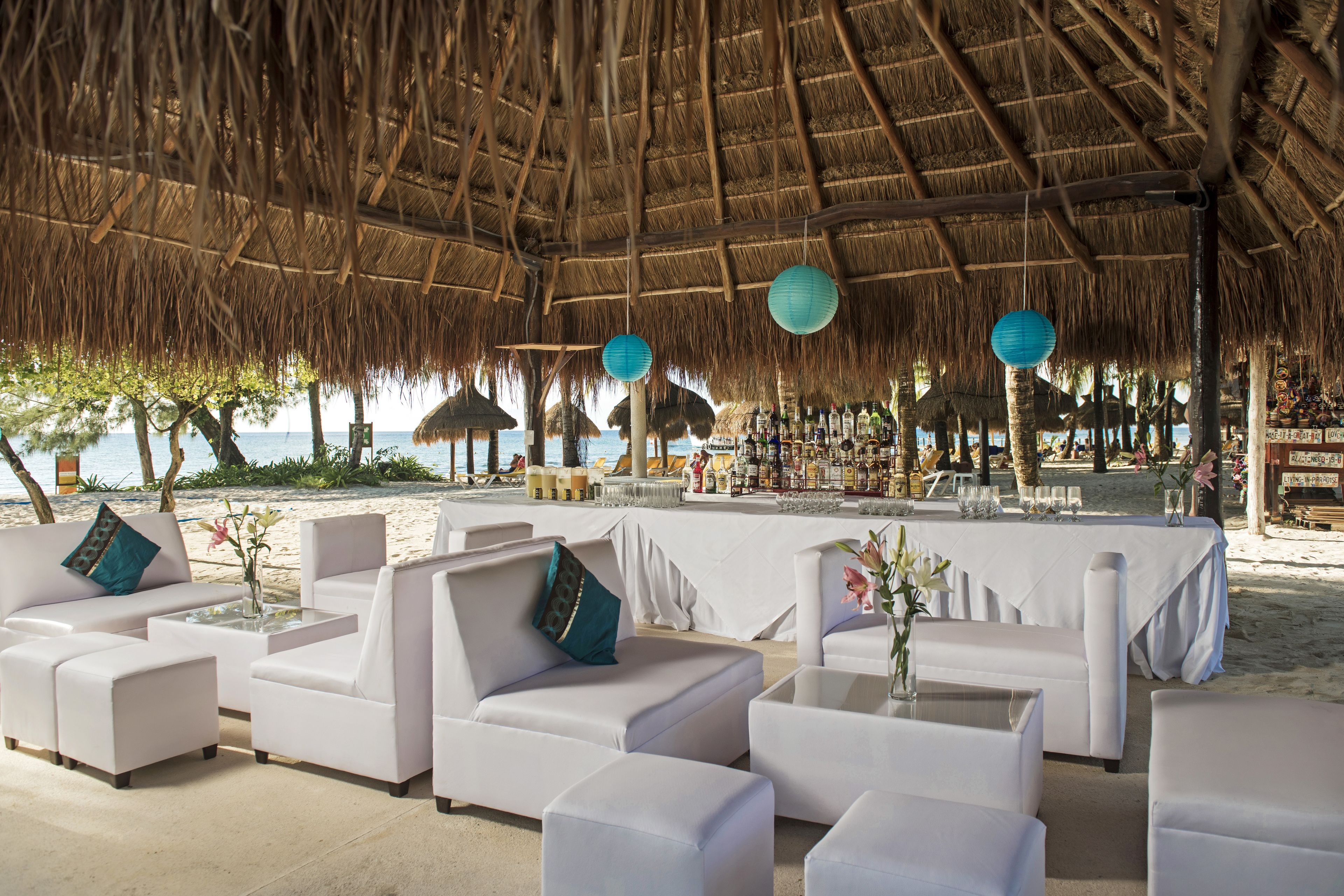 A beachside lounge with white sofas and tables, a thatched roof, and a view of the sea.