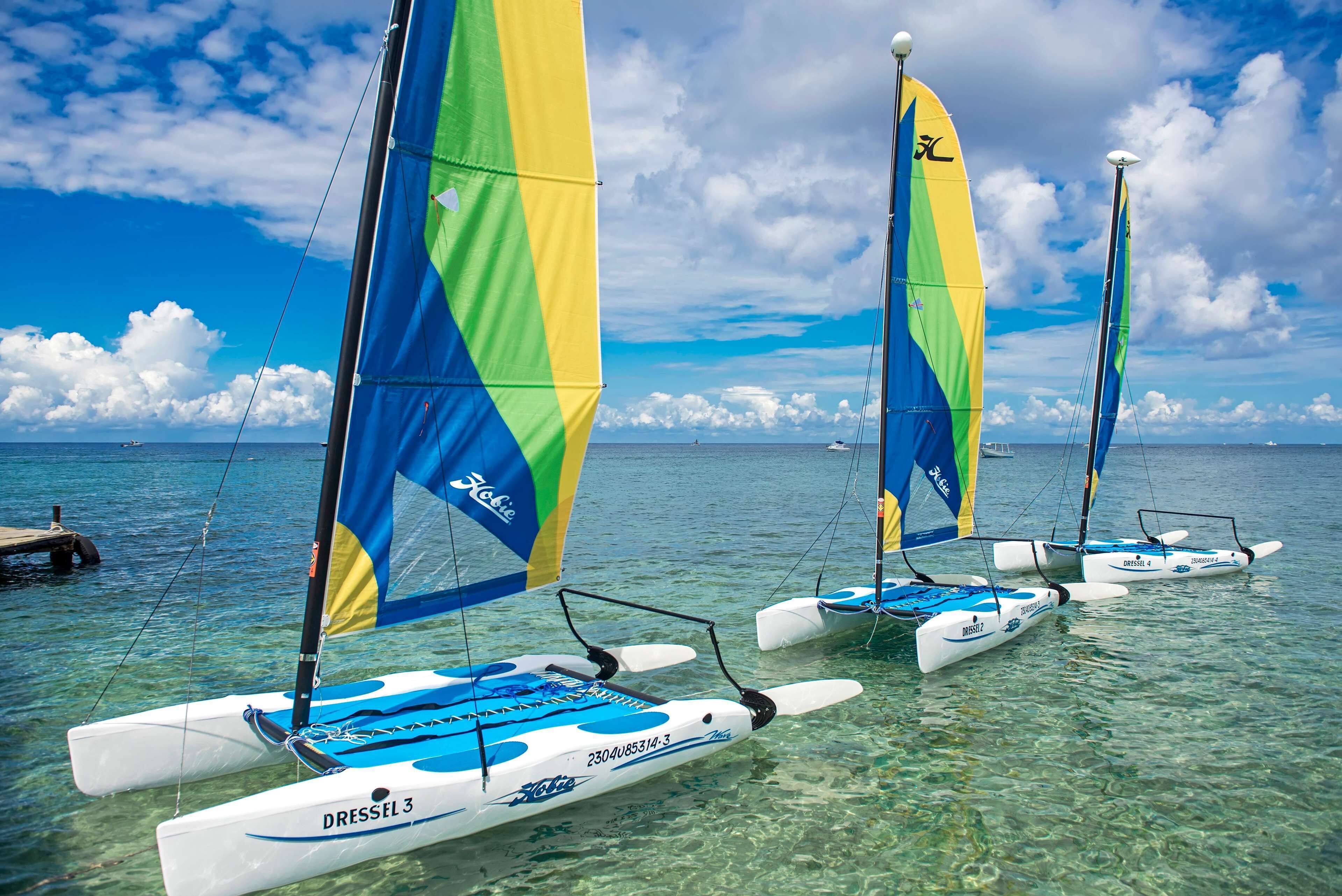 A row of sailboats docked at a marina with clear blue water and a partly cloudy sky.