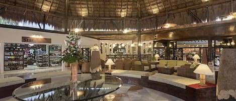 A spacious lobby with a high thatched ceiling, a central glass coffee table, and comfortable seating areas.