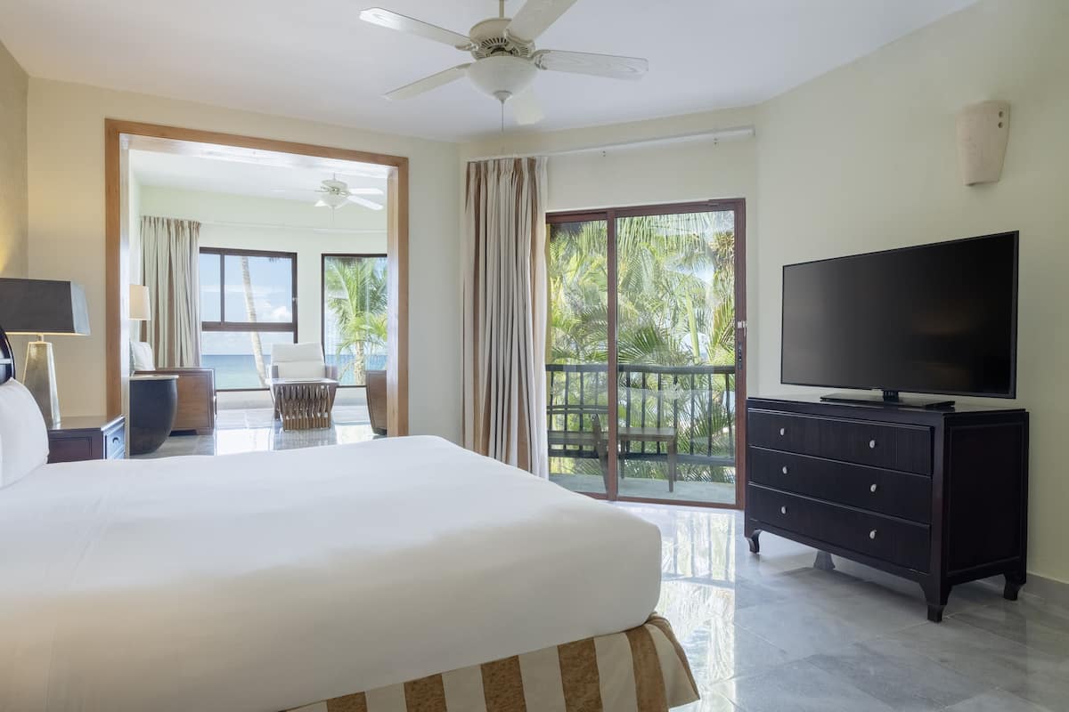 Ein Hotelzimmer mit einem großen Bett, einem Fernseher, einer Kommode und einem Balkon mit Blick auf Palmen.