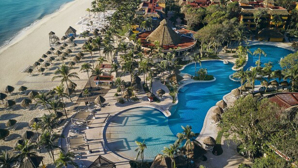 Vista aérea de un complejo turístico con una piscina grande, playa y palmeras.