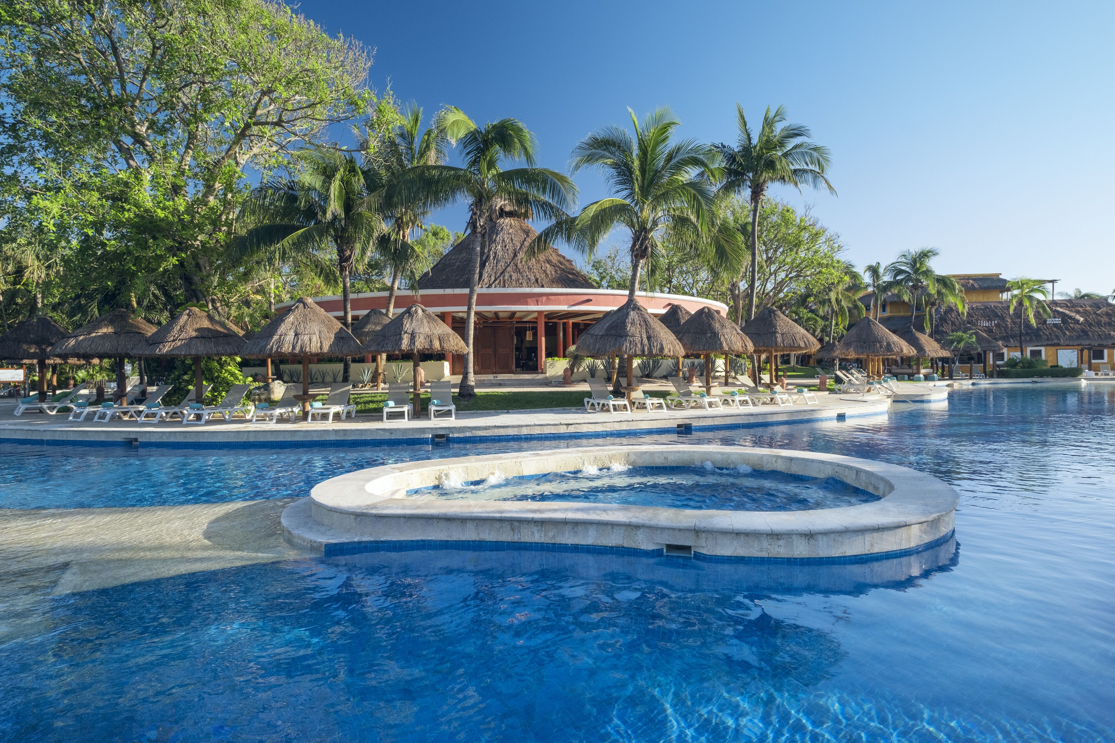 A resort pool area with lounge chairs and thatched umbrellas, surrounded by palm trees and a building with a thatched roof.