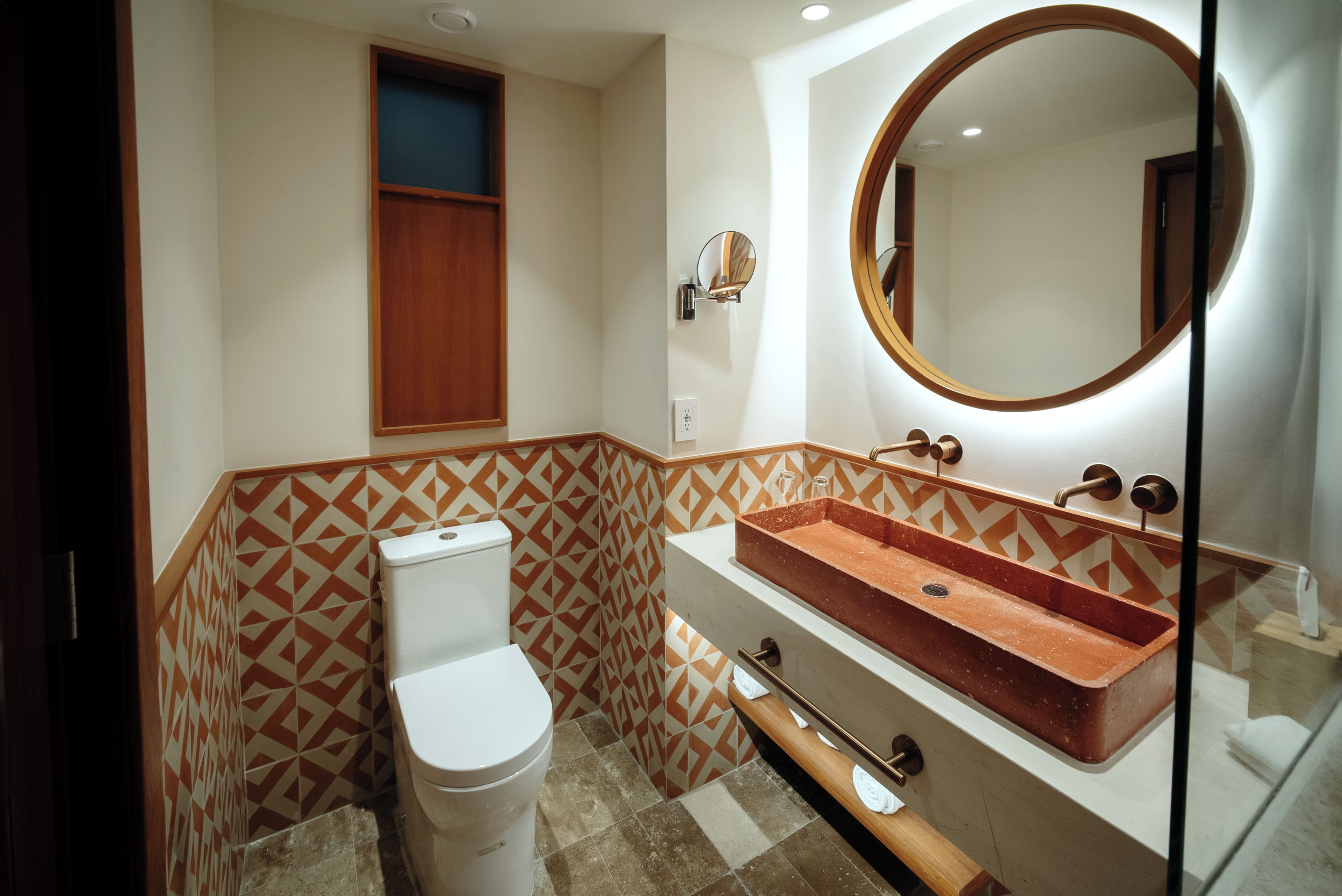 Une salle de bain comprenant des toilettes, un lavabo rectangulaire avec un plan de travail en bois, un miroir fixé au mur et des murs recouverts de carreaux à motifs.