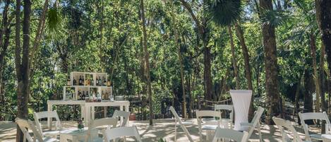 An outdoor seating area with white tables and chairs, surrounded by lush greenery and trees.