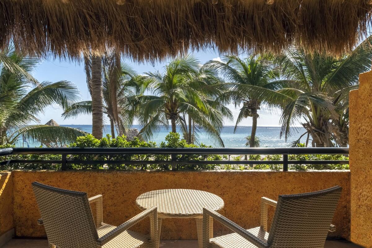 A balcony with wicker furniture overlooking a beach with palm trees and the ocean.