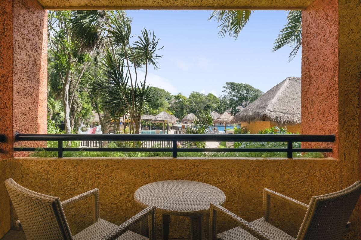 A balcony with wicker chairs and a table, overlooking a pool area with thatched-roof structures and lush greenery.