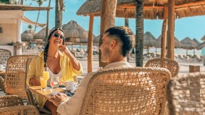 Una pareja cenando en un restaurante frente al mar, con sillas de mimbre y sombrillas de paja.