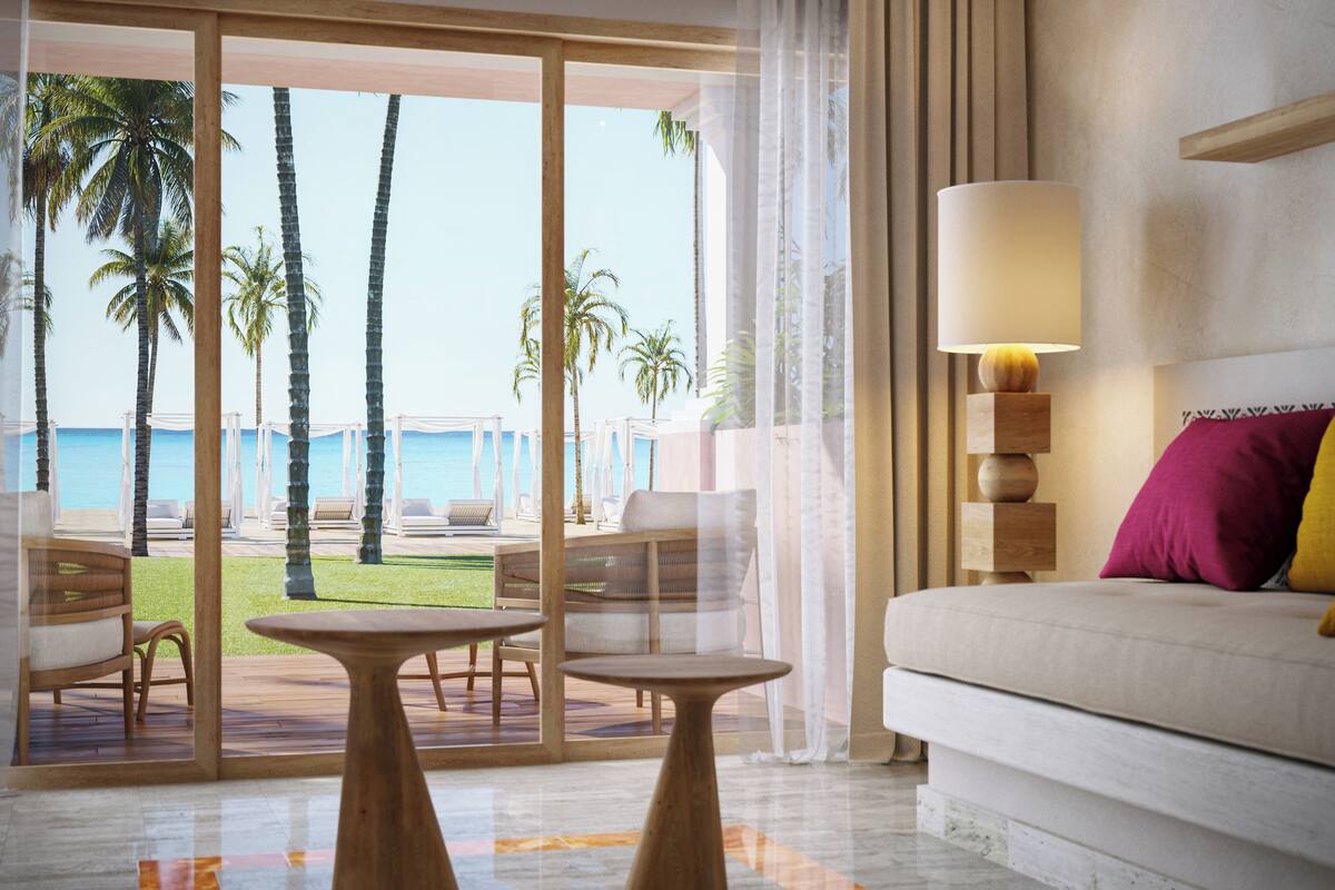 A hotel room with a view of palm trees and the ocean, a sofa, a table, and a lamp.
