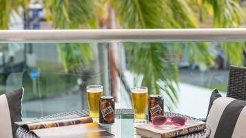 A round table with a glass top, two cans of beer, a glass of beer, and a book titled "Sir Emile Straker" on a balcony overlooking palm trees.