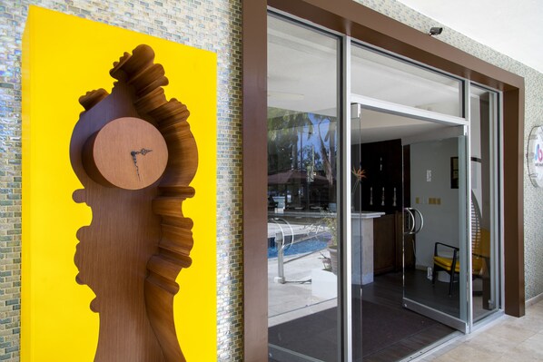 A modern building entrance with a large wooden sculpture of a human head and a clock, a glass door, and a visible pool area. - The Rockley by Ocean Hotels (Rockley)