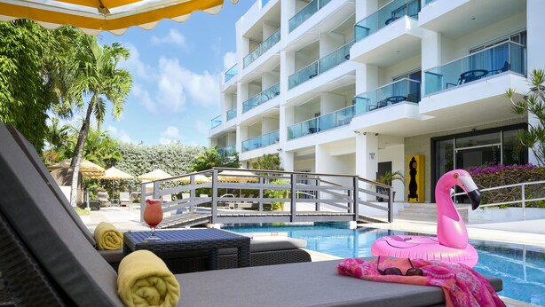 Zona junto a la piscina con sillones de descanso, un flotador con forma de flamenco rosa y un edificio moderno con balcones al fondo.