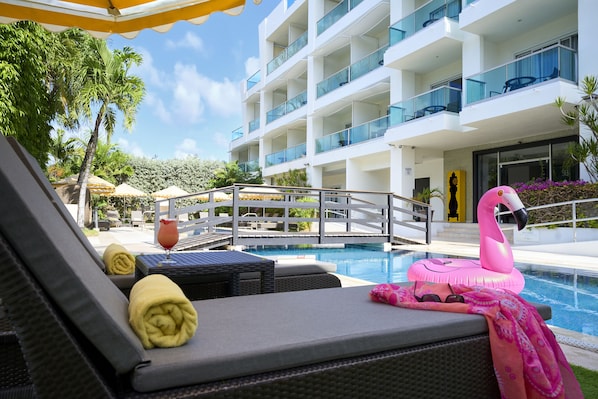 A poolside area with lounge chairs, a pink flamingo float, and a modern building with balconies in the background. - The Rockley by Ocean Hotels (Rockley)