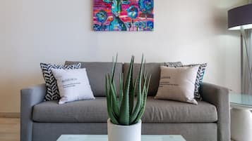 Sala de estar con un sofá gris, dos cojines estampados, una planta de aloe vera en maceta blanca y un cuadro abstracto colorido en la pared.
