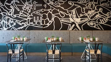 A dining area with a black and white mural, teal cushioned seating, and tables with chairs and small plants.