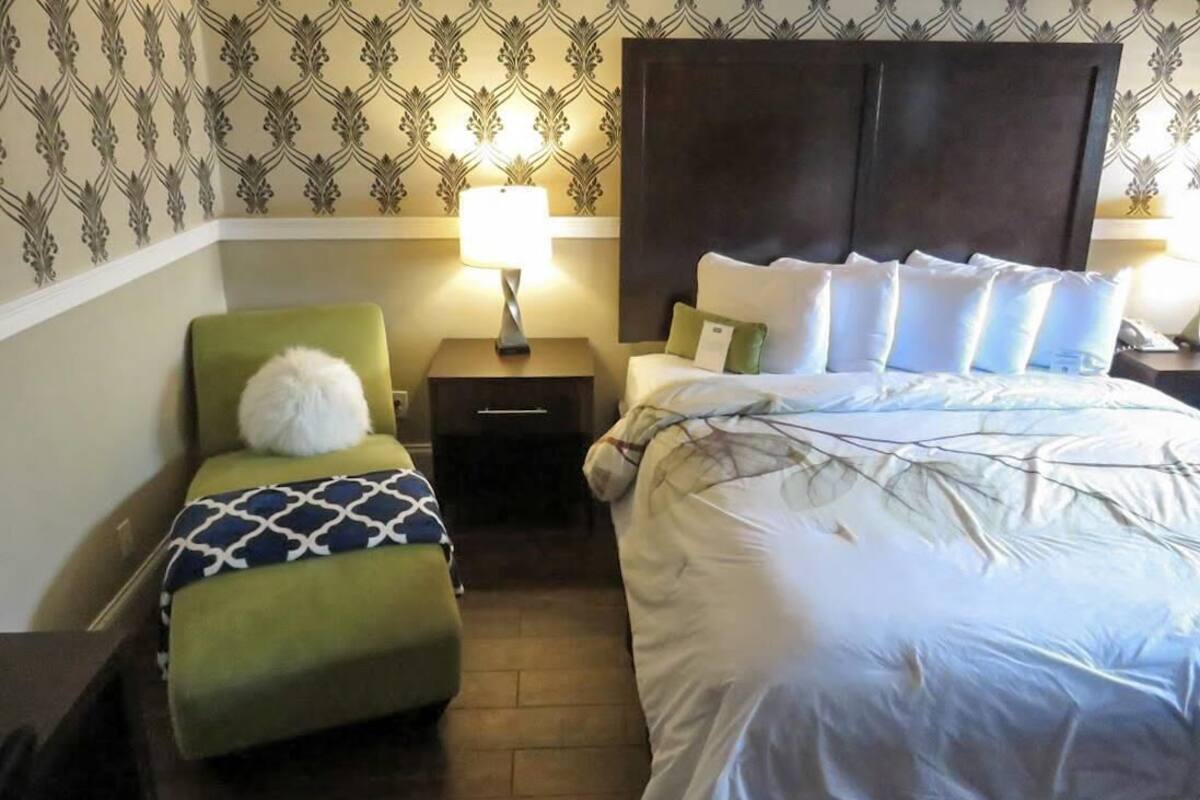 A hotel room with a large bed, a green armchair, a nightstand with a lamp, and patterned wallpaper.