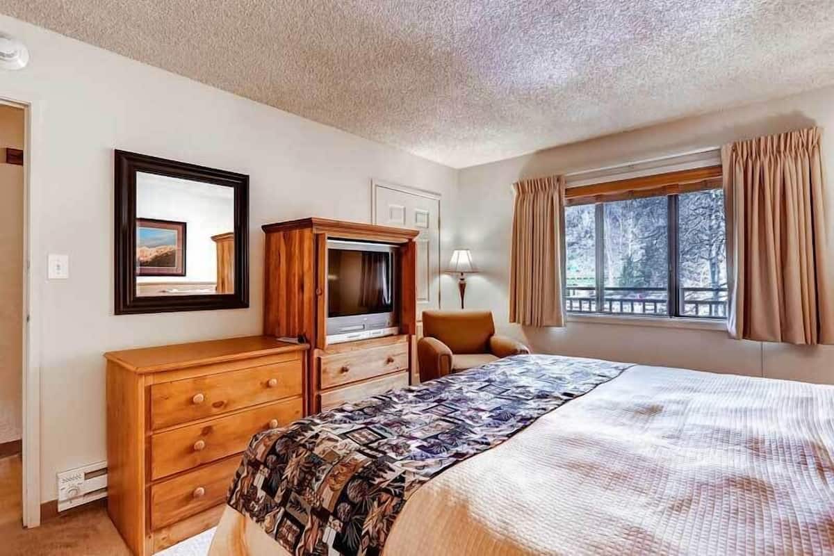 Un dormitorio con cama, cómoda de madera, televisor, silla y ventana con cortinas.
