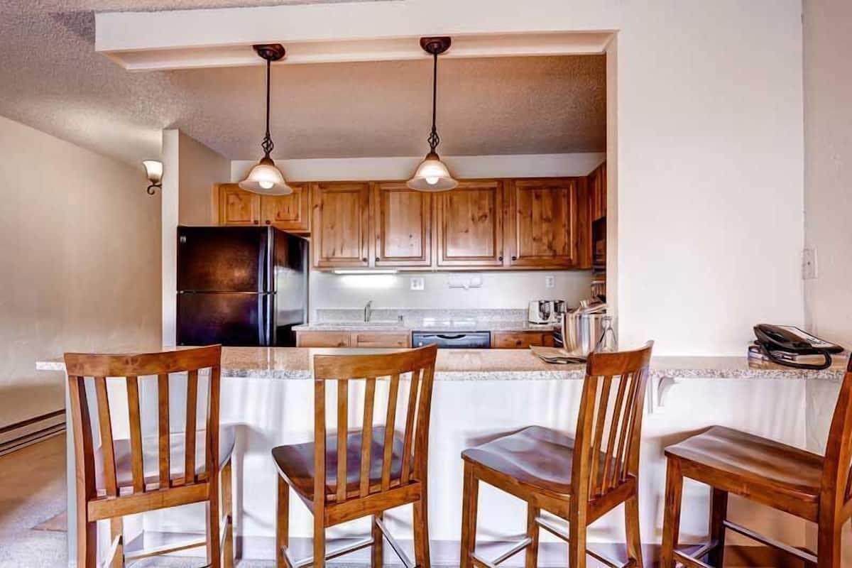 Una cocina con armarios de madera, un refrigerador, un lavamanos y una zona de comedor con cuatro sillas de madera.