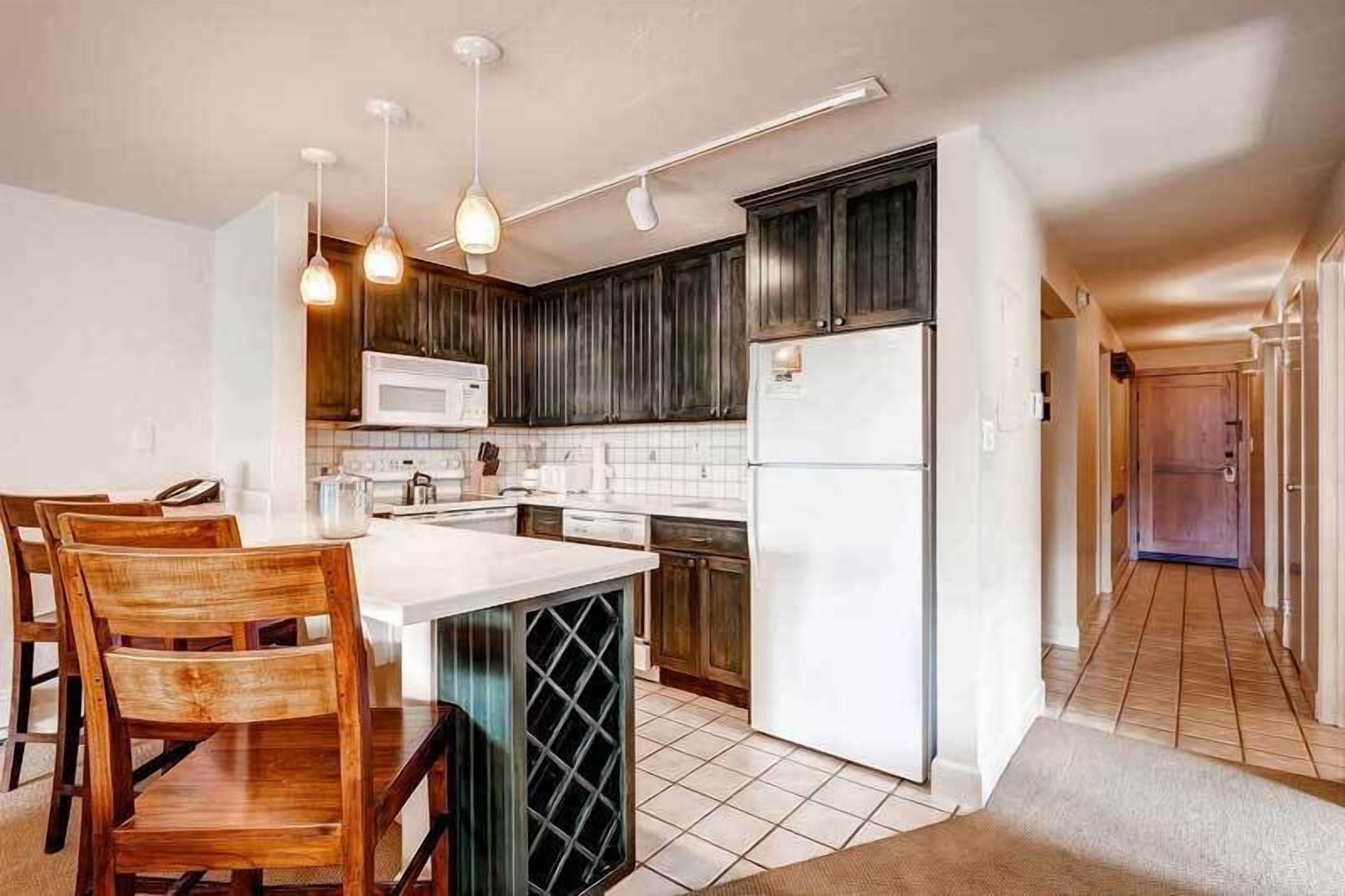 A modern kitchen with dark wooden cabinets, a white island with bar stools, a built-in wine cooler, and a stainless steel refrigerator.