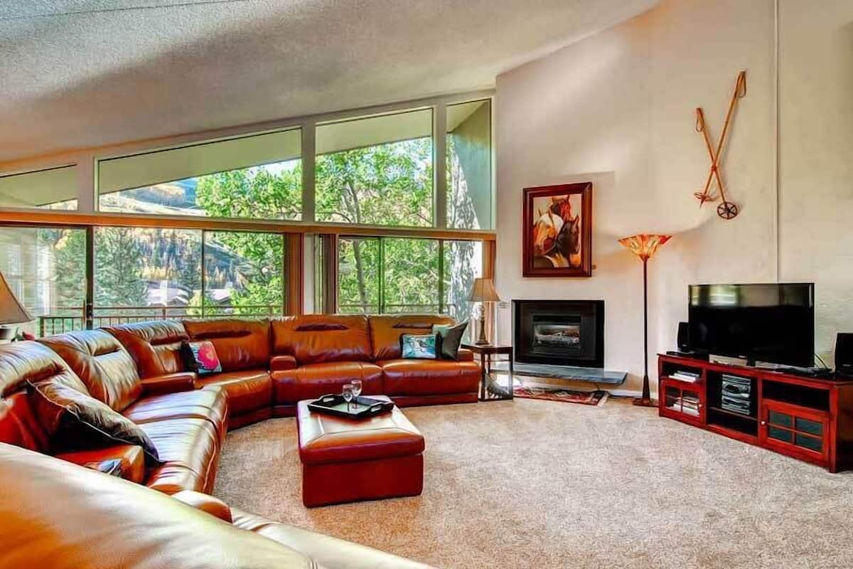 Una sala de estar moderna con un sofá de cuero tipo seccional, una chimenea, un televisor y ventanas amplias con vistas a la vegetación.
