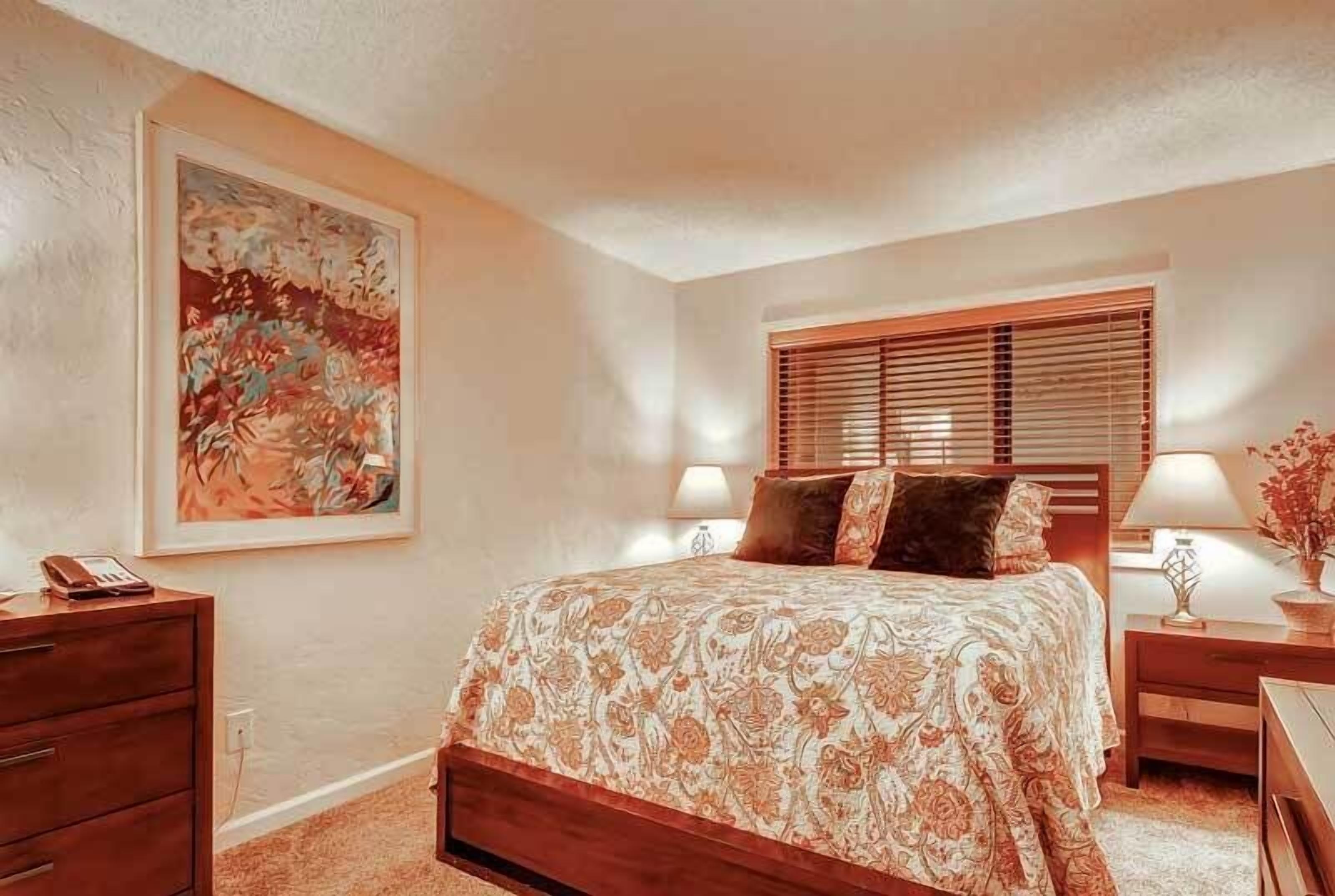 Une chambre à coucher avec un grand lit, deux tables de chevet avec des lampes, une commode en bois et un grand tableau floral accroché au mur.