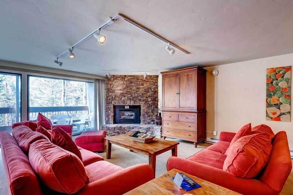 Una sala de estar moderna con chimenea de piedra, muebles rojos y un ventanal con vistas a árboles.