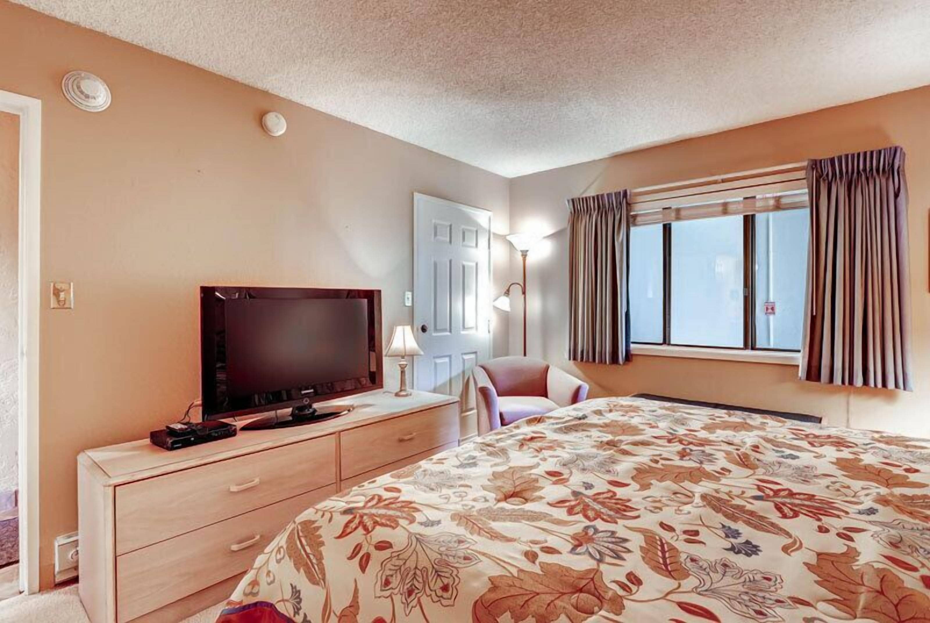 A hotel room with a bed, a TV on a wooden dresser, a door, and a window with curtains.