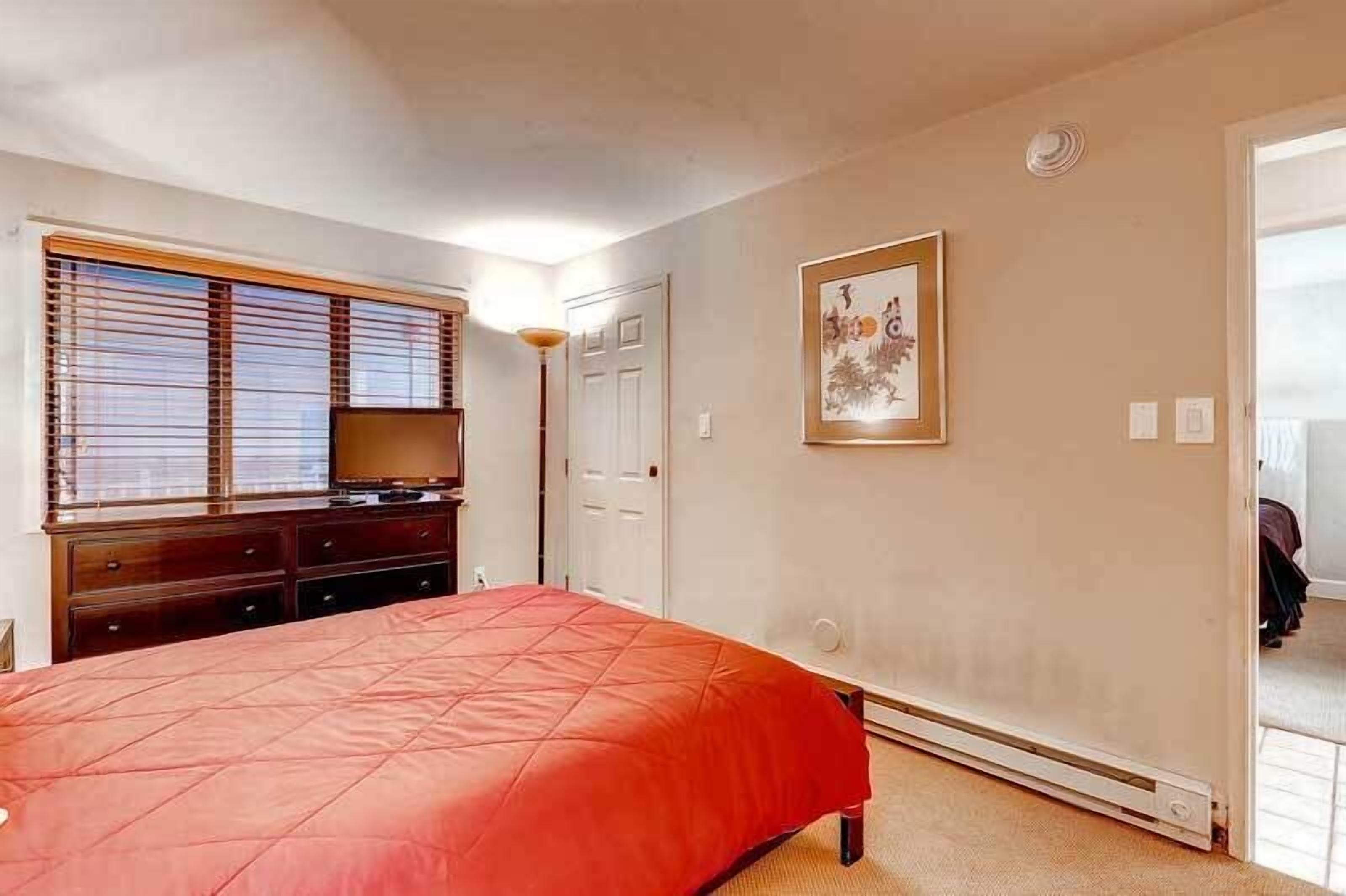 Ein Schlafzimmer mit Bett, Kommode, Rollläden, einer Tür und einem gerahmten Bild an der Wand.
