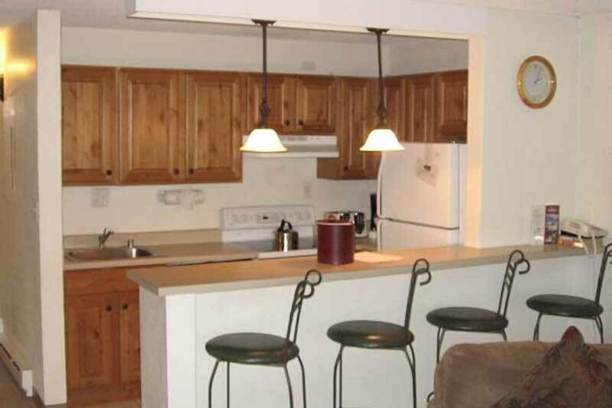 Una cocina con armarios de madera, una isla central con taburetes de bar, iluminación colgante y un reloj en la pared.