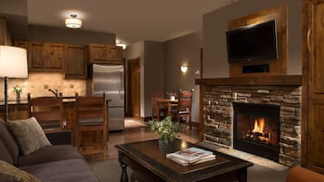 Ein Wohnzimmer mit Kamin, einem darüber montierten Fernseher, Holzmöbeln und einer Küchenzeile im Hintergrund.