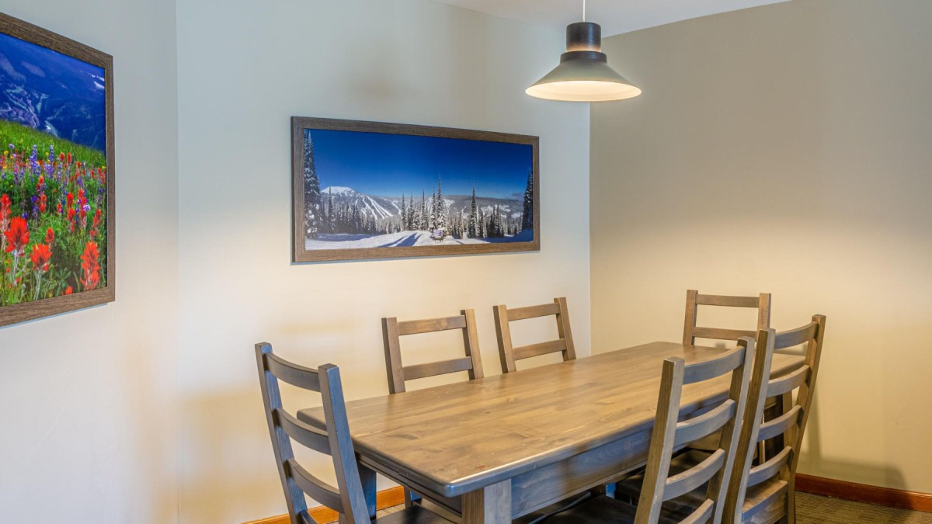Un área de comedor con una mesa y sillas de madera, un cuadro de un paisaje montañoso nevado y una lámpara colgante.