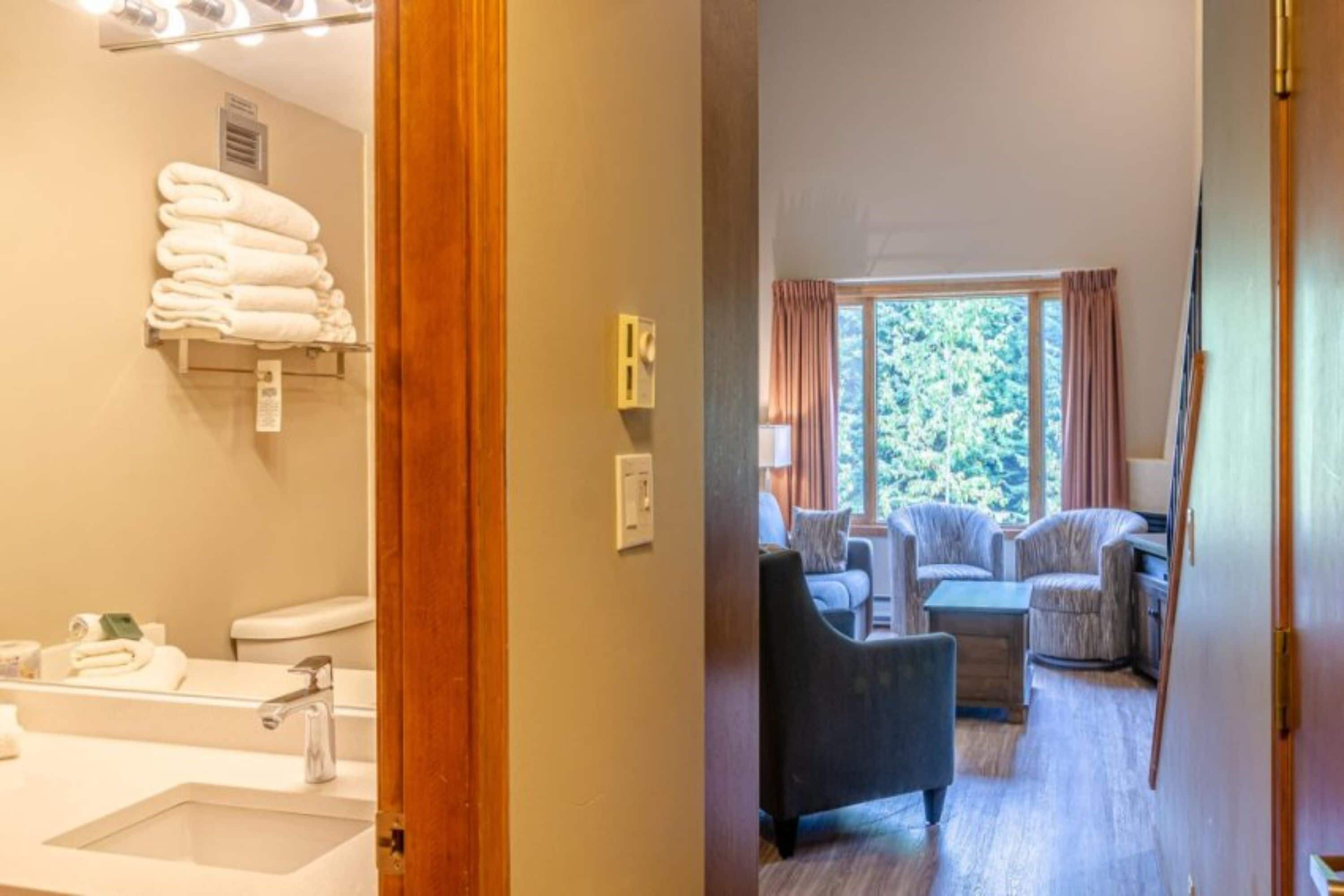 A bathroom with a sink, toilet, and towels. There is a window with curtains and a view of greenery.