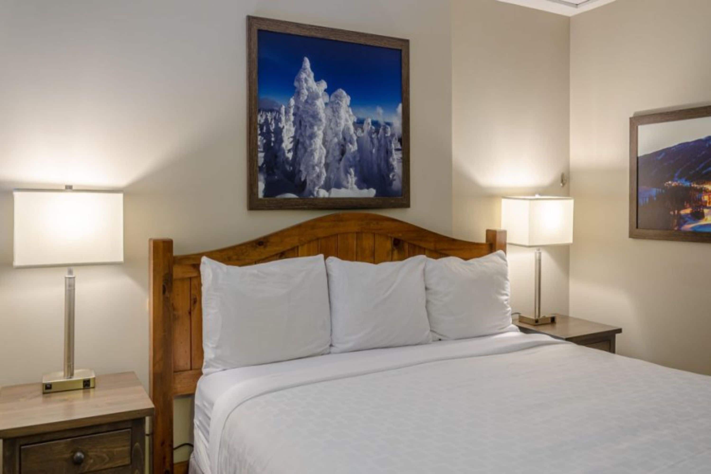 Hotelkamer met een houten hoofdbord, wit beddengoed, twee bedlampen en een lijst met een afbeelding van een besneeuwd berglandschap aan de muur.
