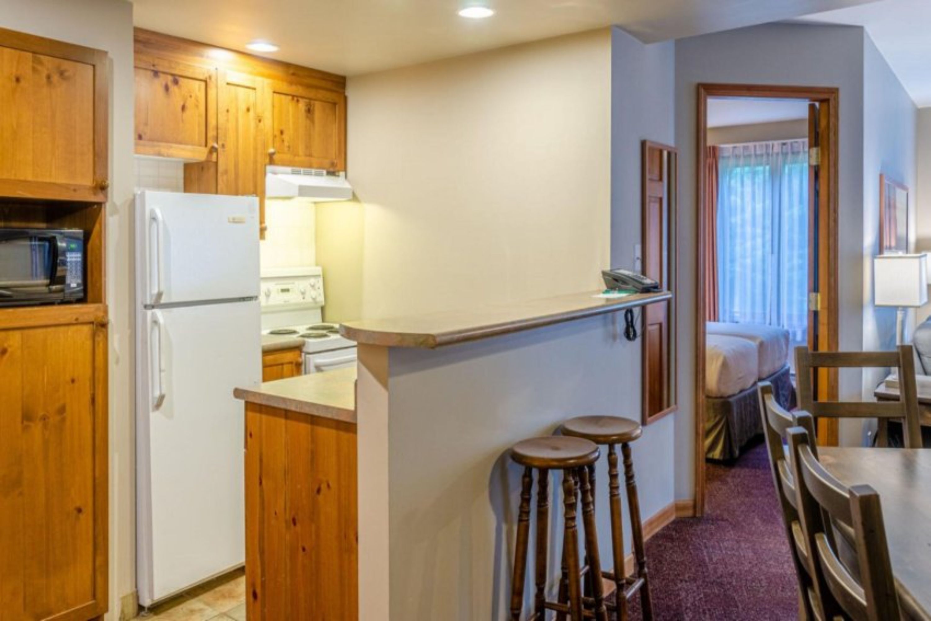A compact kitchen with wooden cabinets, a microwave, a refrigerator, and a small dining area with stools.