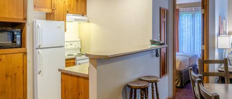 A compact kitchen with wooden cabinets, a microwave, a refrigerator, and a small dining area with stools.