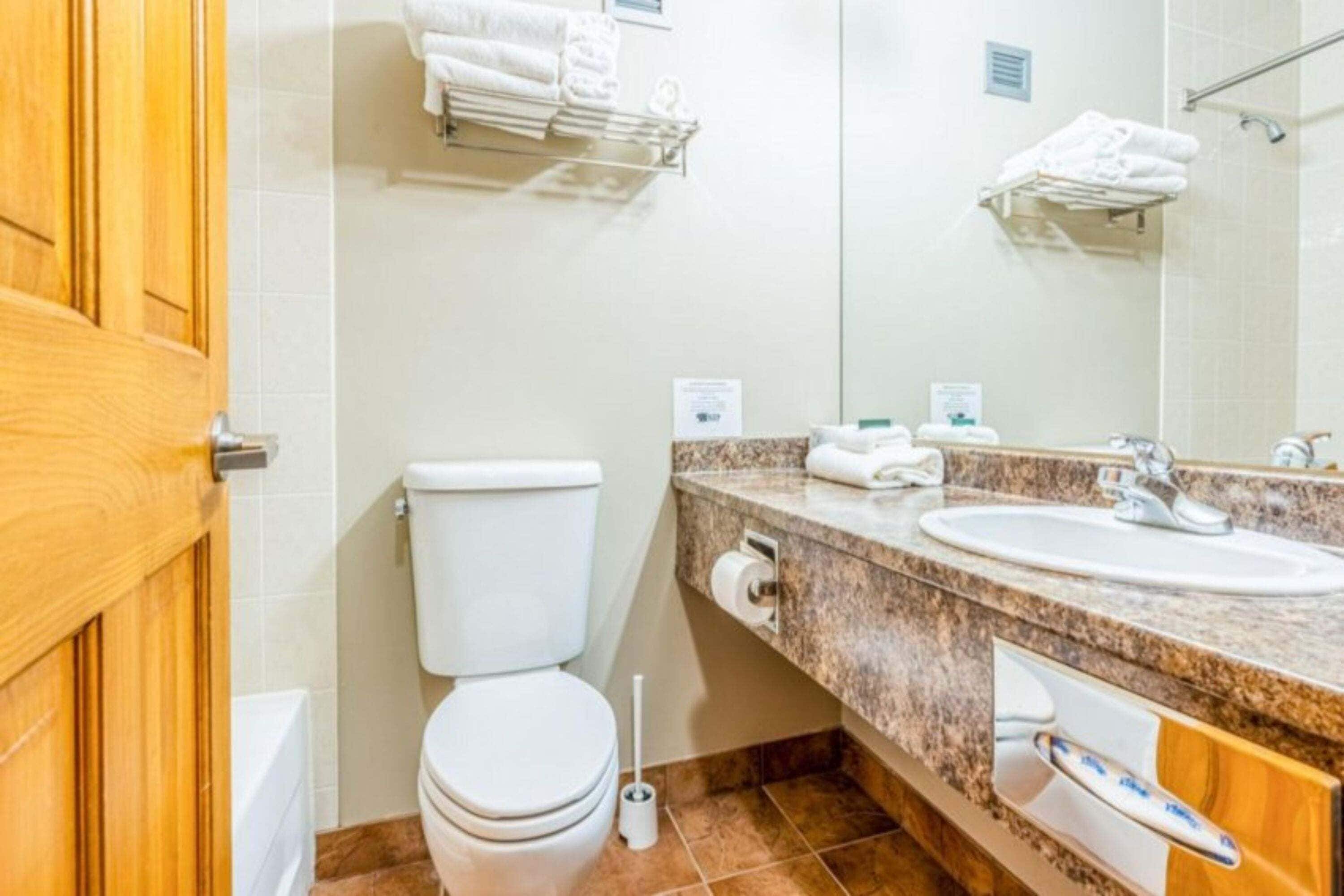 Un baño con inodoro, lavamanos, espejo, toallero y puerta de madera.