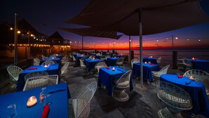Un área de comedor al aire libre con mesas preparadas para dos, cubiertas con manteles azules y sillas de alambre blancas, con vistas a un atardecer sobre el agua.