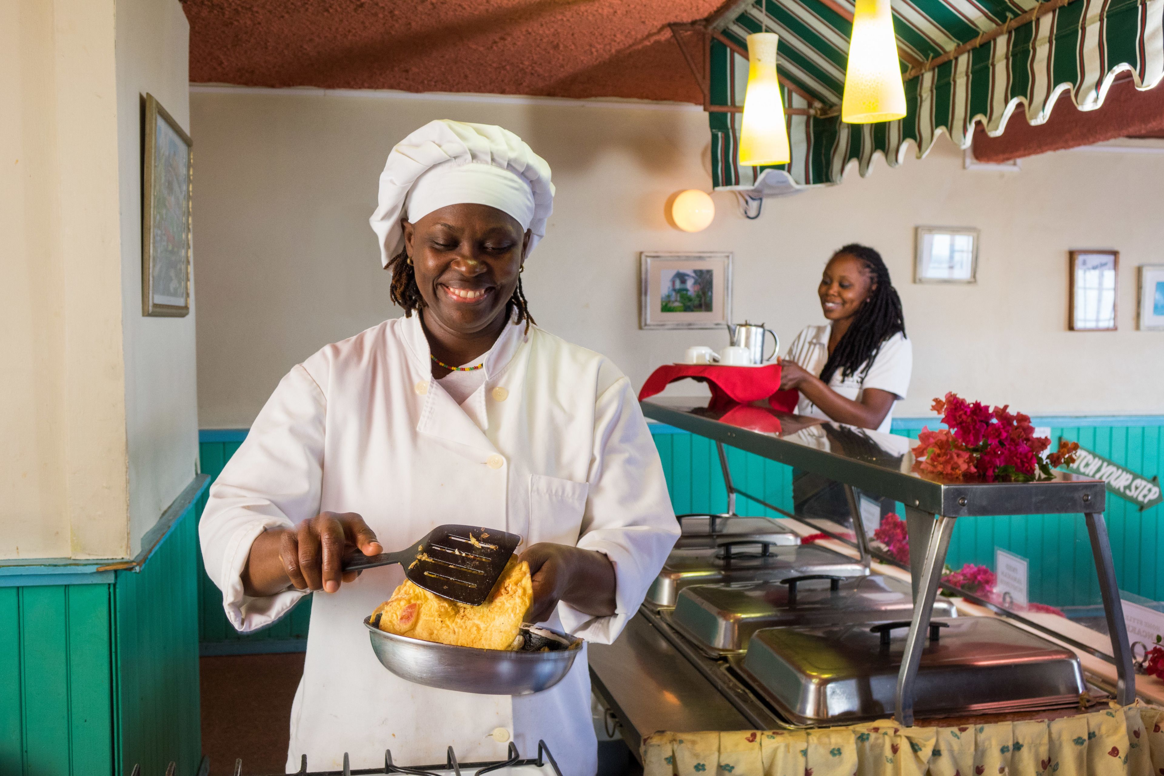 A chef in a white uniform holding a pan with a dish, in a restaurant with a counter and another person in the background.
