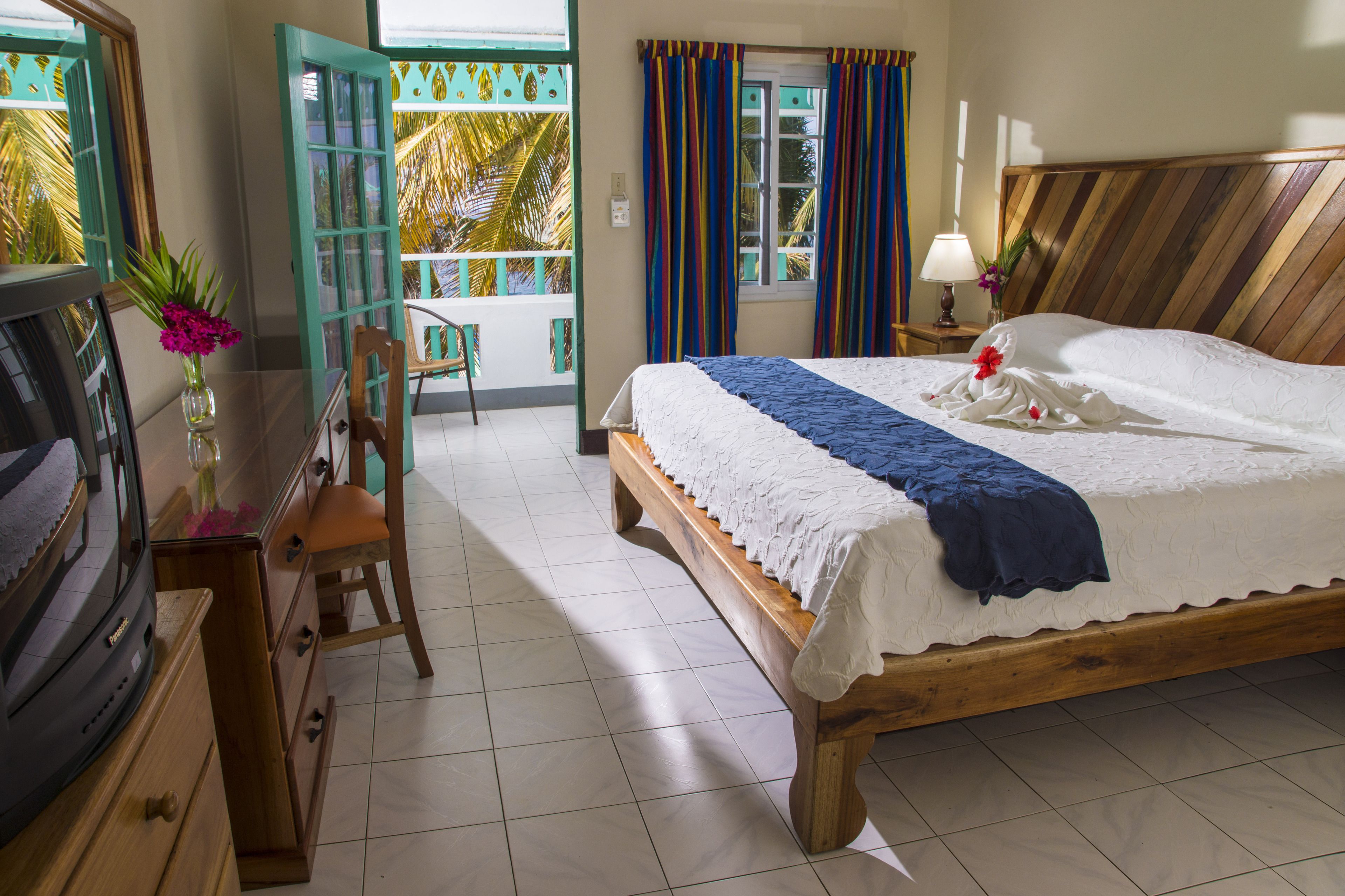 Quarto de hotel com cama de madeira, escrivaninha com televisão, cadeira e vista para palmeiras pela porta aberta.