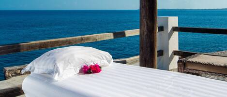 Una cama con ropa blanca y una flor rosa, con vistas a un mar azul cristalino.