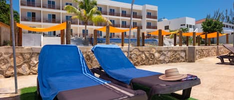 A poolside area with sun loungers covered with blue towels, a hat on one of the loungers, and an orange canopy overhead.