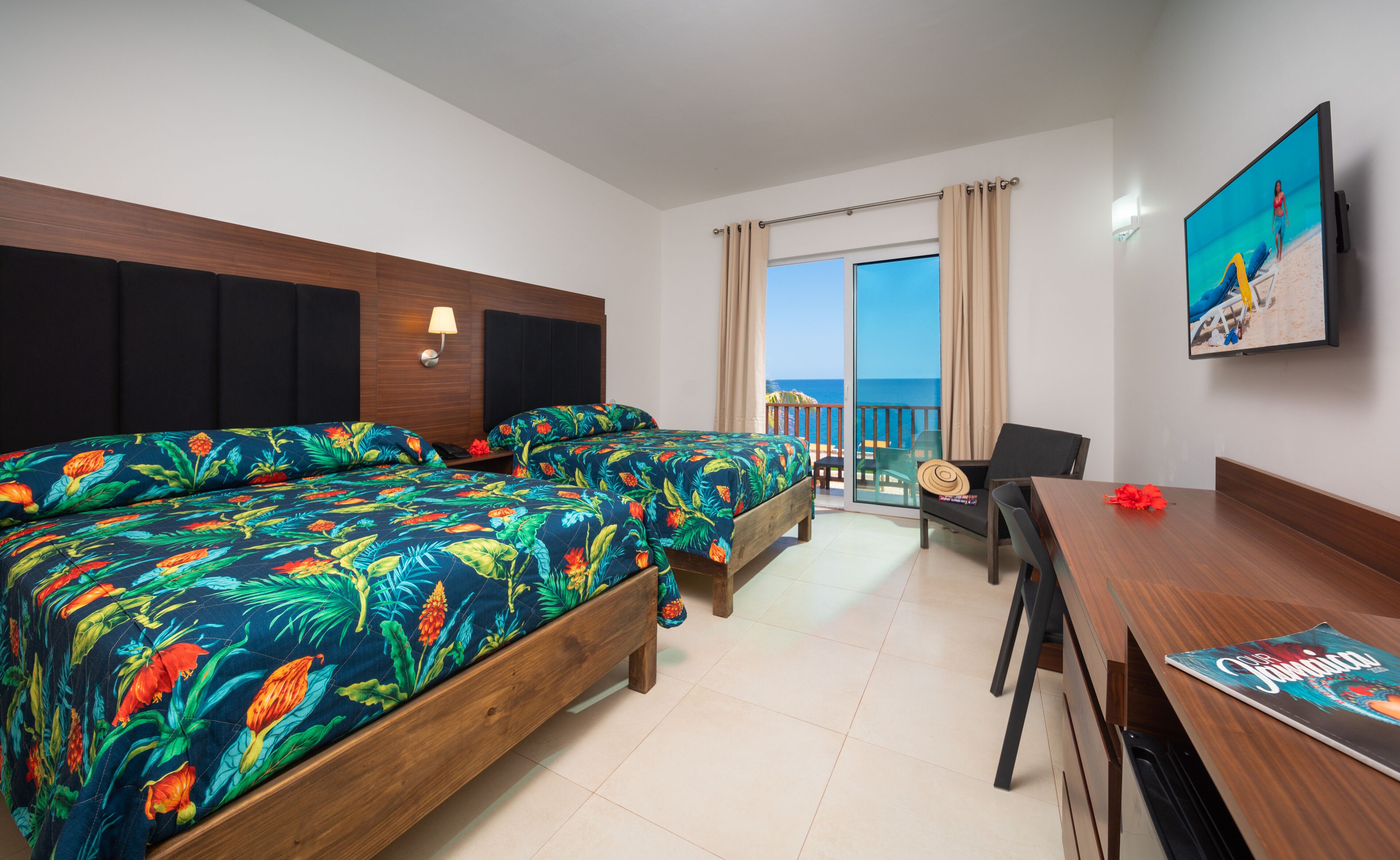 Habitación de hotel con dos camas, un balcón con vista al mar, un escritorio y un televisor en la pared.